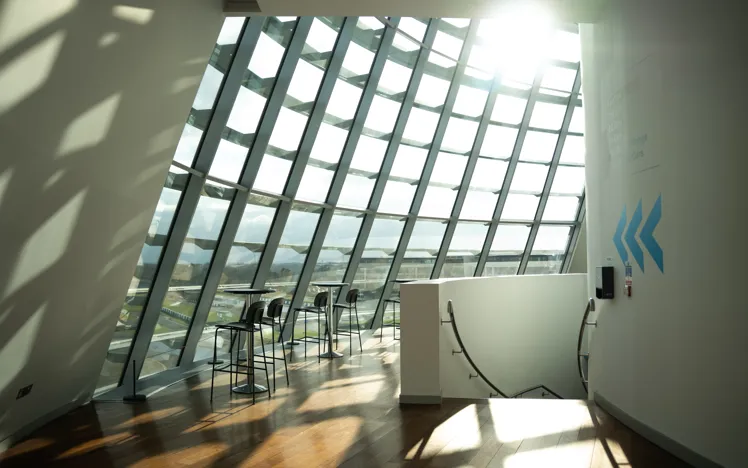 Sunlight streams through the large angled glass windows of a modern building, illuminating a seating area with tall chairs and tables overlooking a track below.