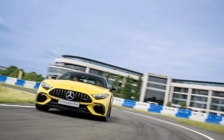 Bright yellow Mercedes-AMG speeding around a bend at Mercedes-Benz World, with driver and passenger enjoying a dynamic track experience in front of a modern building backdrop.
