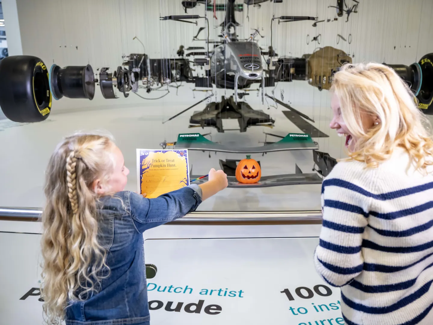A young girl and woman smiling while viewing a suspended Formula 1 car display with a Halloween pumpkin decoration and a "Trick or Treat Pumpkin Hunt" flyer.
