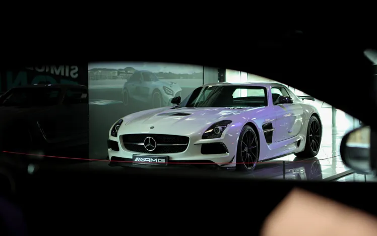 A white Mercedes-AMG sports car is displayed in a sleek showroom with polished floors, large windows, and a branded AMG display wall in the background.