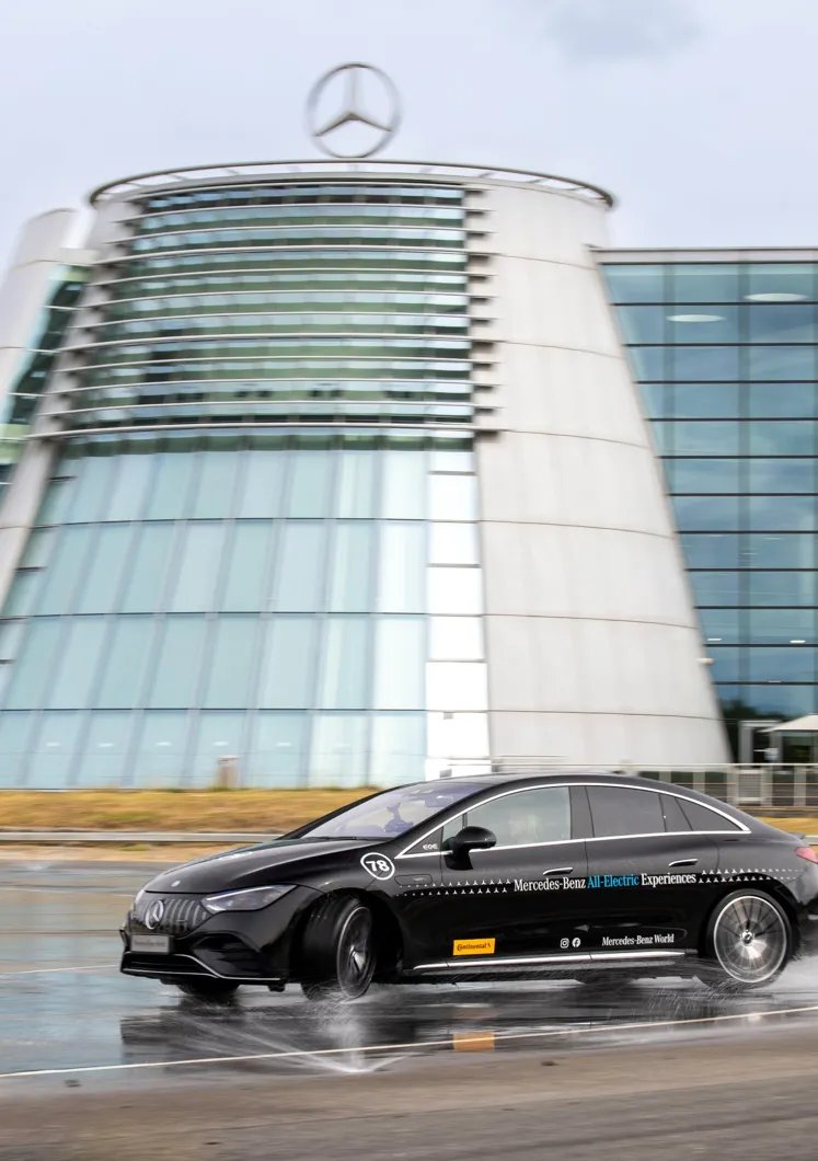 A black Mercedes-Benz EQE electric car drives through water on a test track in front of the iconic glass-fronted Mercedes-Benz World building.