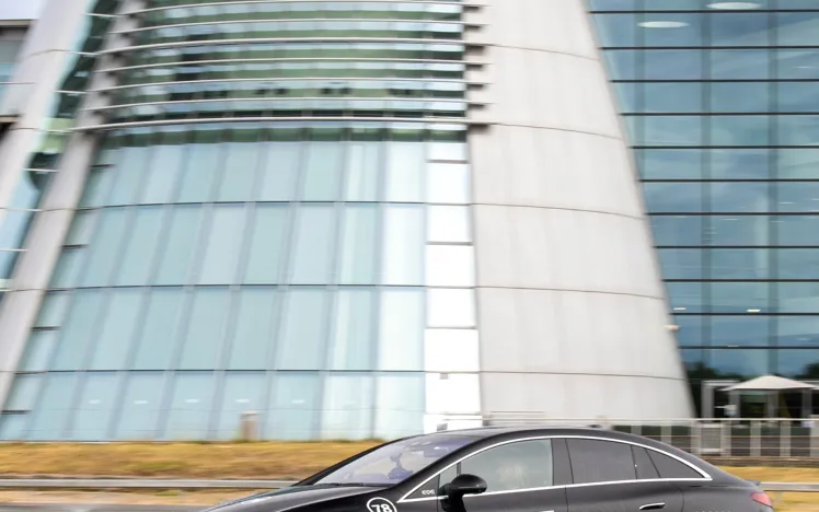 A black Mercedes-Benz EQE electric car drives through water on a test track in front of the iconic glass-fronted Mercedes-Benz World building.