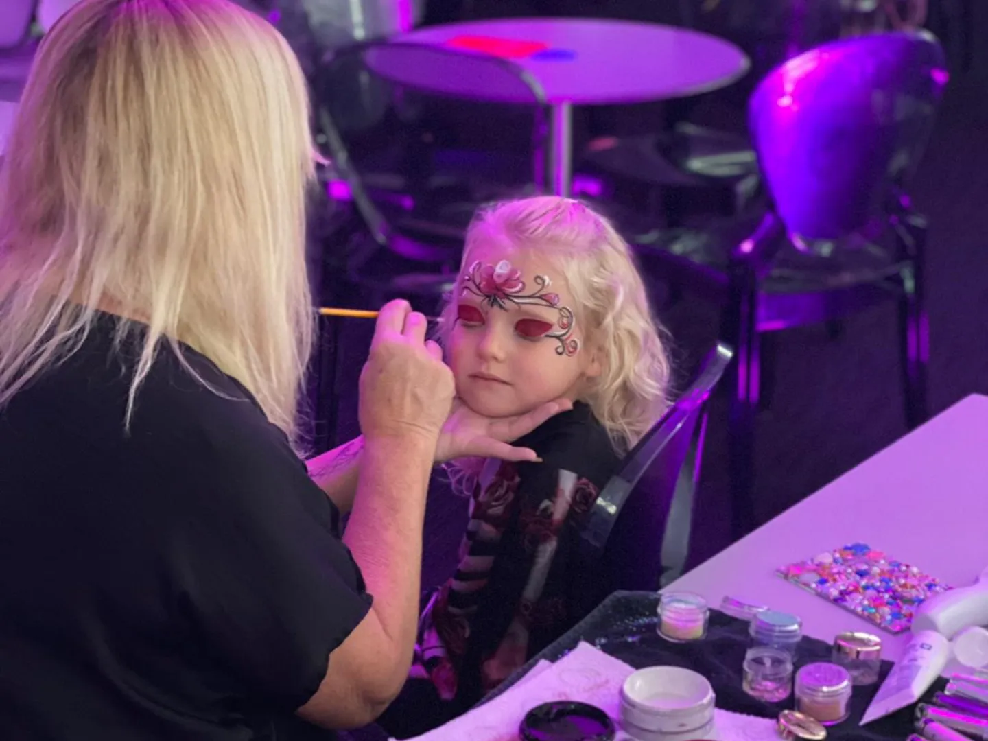 A young girl with blonde hair gets her face painted with a floral design by a woman at a face painting station, surrounded by colorful paints and brushes in a dimly lit room.