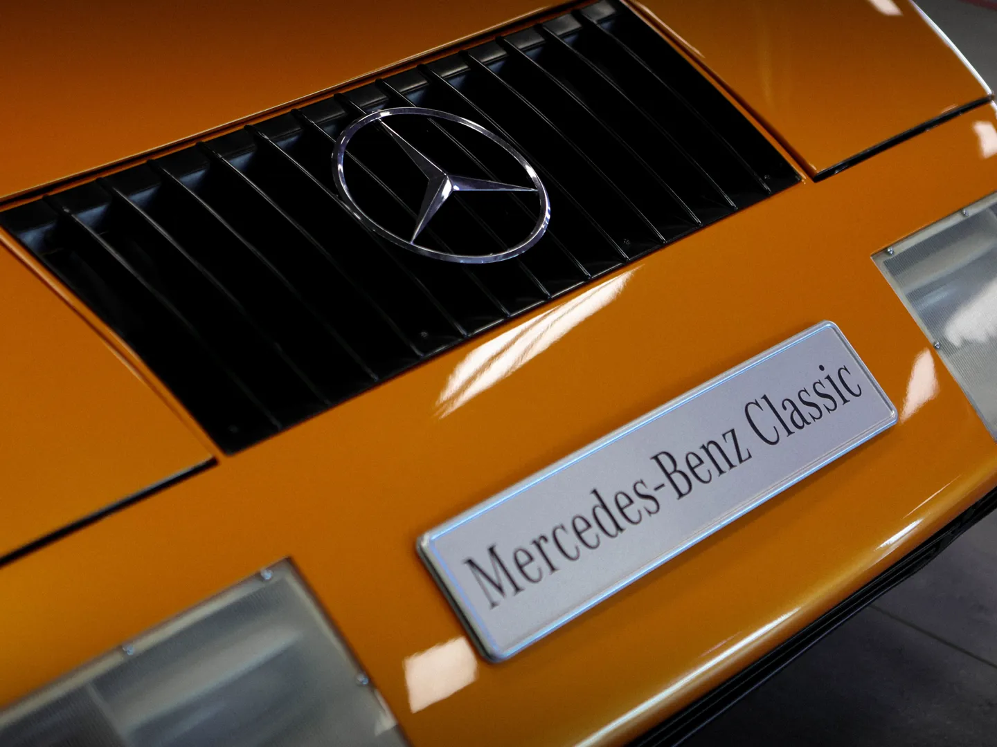 Close-up of a classic Mercedes-Benz vehicle with a bright orange exterior, featuring the iconic grille and a "Mercedes-Benz Classic" nameplate.